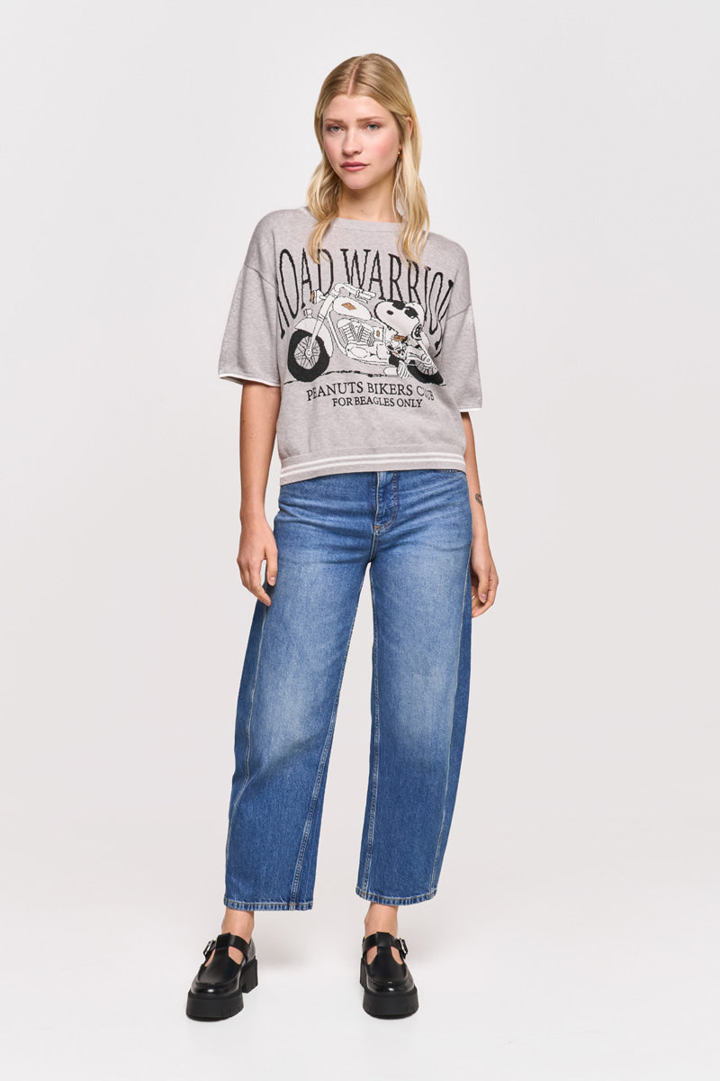 Eine blonde Frau trägt ein graues Princess Goes Hollywood Road Warrior-Grafik-T-Shirt mit einem Cartoon-Biker, blaue Jeans mit weitem Bein und schwarze Plateausandalen. Sie steht vor einem weißen Hintergrund und posiert entspannt für die Kamera.