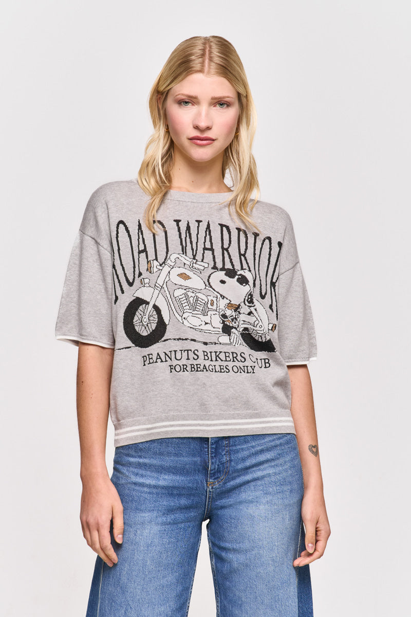 Eine Frau mit langen blonden Haaren trägt ein graues Princess Goes Hollywood-T-Shirt mit Snoopy auf einem Motorrad und der Aufschrift "ROAD WARRIOR-Peanuts Bikers Club for Beagles Only", gepaart mit blauen Jeans, vor einem weißen Hintergrund.