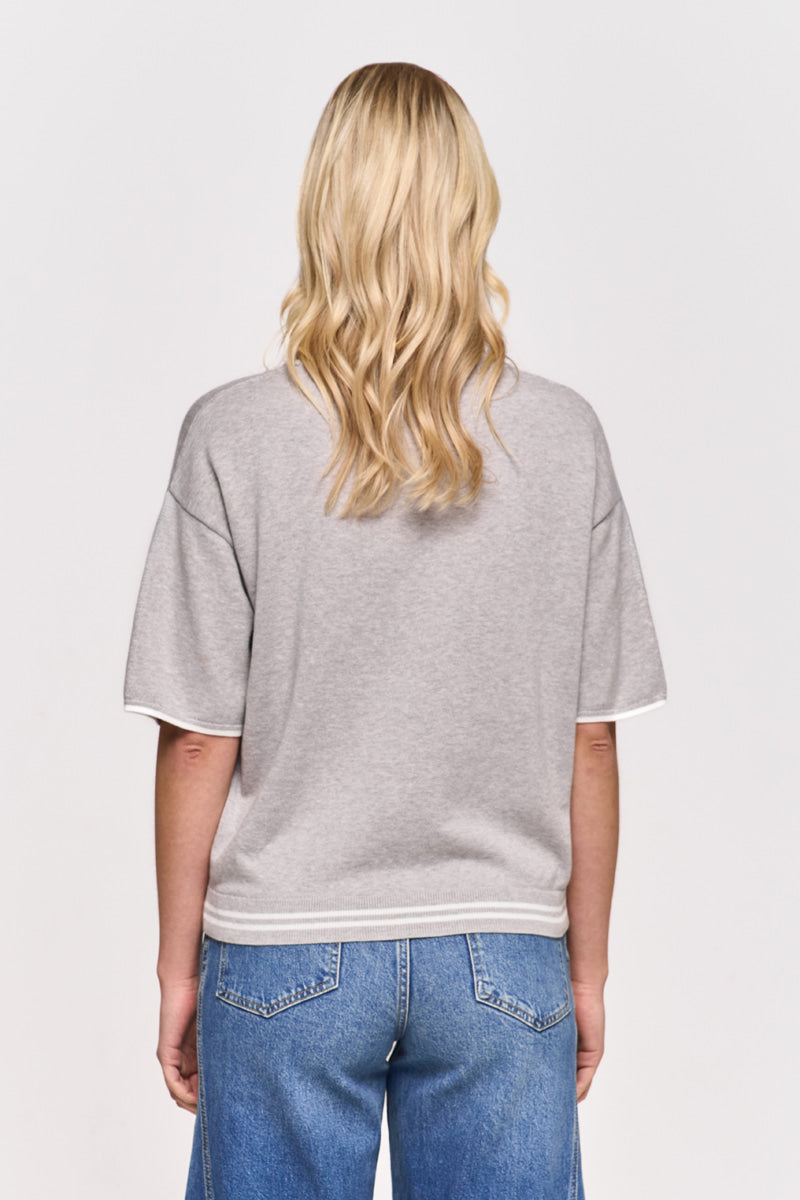 Eine Person mit gewelltem blondem Haar wird von hinten gezeigt. Sie trägt ein lockeres graues Princess Goes Hollywood-T-Shirt mit kurzen Ärmeln und weißem Saum sowie blaue Jeans. Der einfache Hintergrund unterstreicht die lockere, entspannte Atmosphäre.