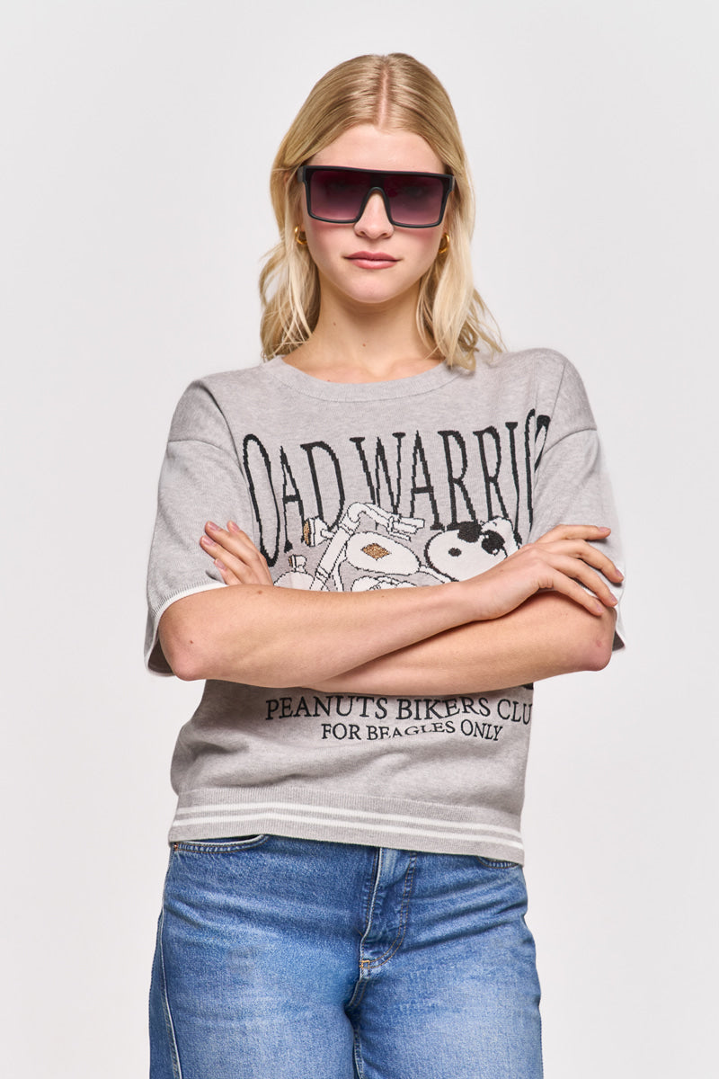 Eine Frau mit langen blonden Haaren trägt eine große, dunkle, rechteckige Sonnenbrille und ein hellgraues Princess Goes Hollywood-T-Shirt mit einer Snoopy-Cartoon-Grafik, auf der "ROAD WARRIOR" und "PEANUTS BIKERS CLUB FOR BEACHES ONLY" zu lesen sind, sowie blaue Jeans.