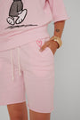 Eine Person trägt eine hellrosa Princess Goes Hollywood-Sweatshorts und ein passendes Oberteil mit einer Cartoon-Handschuh-Grafik. Die weichen Baumwollshorts haben einen Kordelzug und ein Herzstickerei-Detail; ihre Hand zeigt ein Herz-Tattoo und ein klobiges Goldarmband.