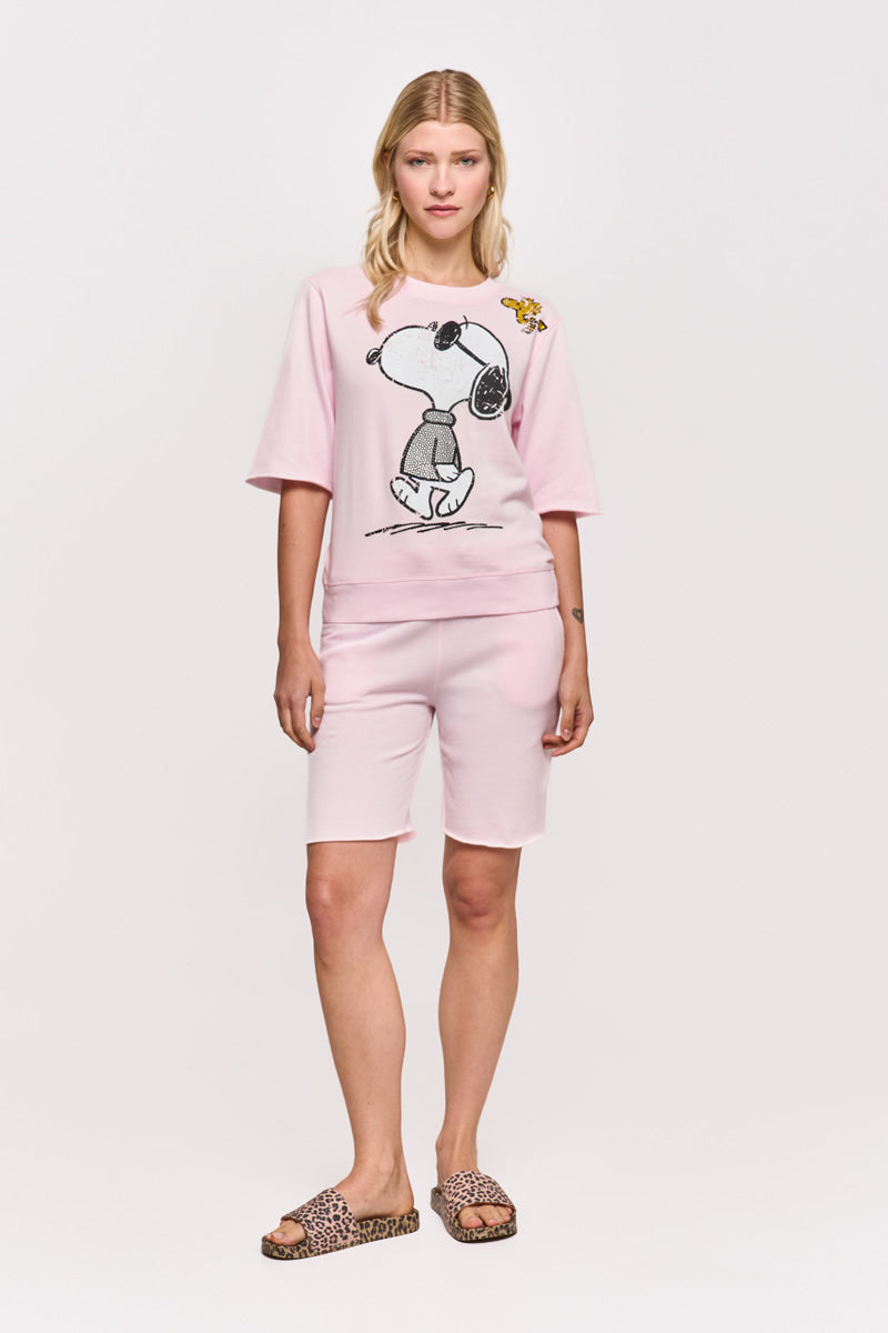 Eine Frau mit blondem Haar und neutralem Gesichtsausdruck blickt nach vorne. Sie trägt ein hellrosa Princess Goes Hollywood Snoopy &amp; Woodstock Baumwolle T-Shirt und knielange Sweatshorts, gepaart mit Slide-Sandalen mit Leopardenmuster vor einem weißen Hintergrund.