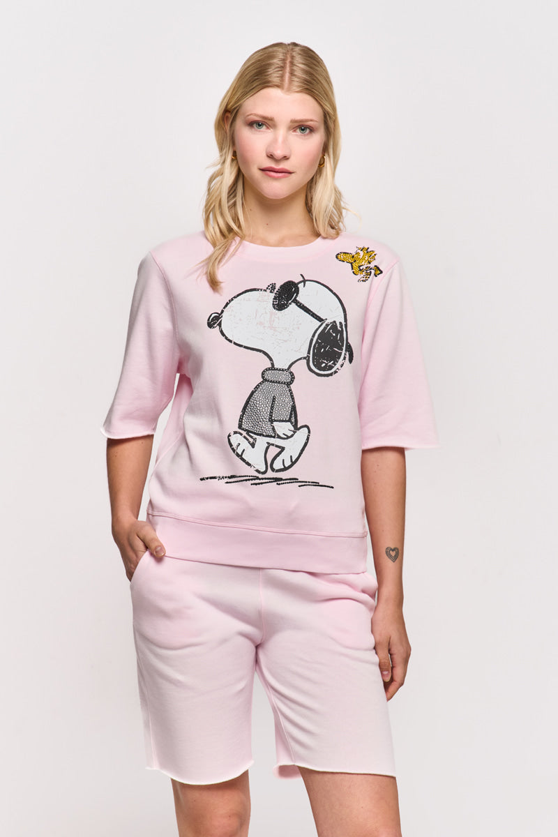 Eine junge blonde Frau modelliert ein hellrosafarbenes Princess Goes Hollywood-Baumwollset - eine Sweathose und ein kurzärmeliges Oberteil mit einer großen Grafik von Snoopy mit Sonnenbrille und Woodstock am Ohr - und posiert selbstbewusst vor einem schlichten Hintergrund.
