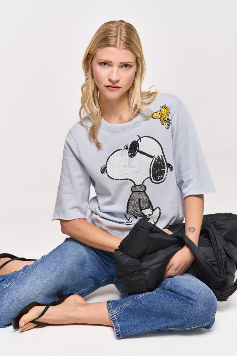 Eine Frau mit langen blonden Haaren sitzt in einem hellgrauen Princess Goes Hollywood Snoopy-Print Sweatpullover, blauen Jeans und schwarzen Riemchensandalen auf dem Boden. Sie hält eine schwarze Puffjacke in der Hand, die ein Herz-Tattoo auf ihrem linken Unterarm zeigt. Der Hintergrund ist weiß.