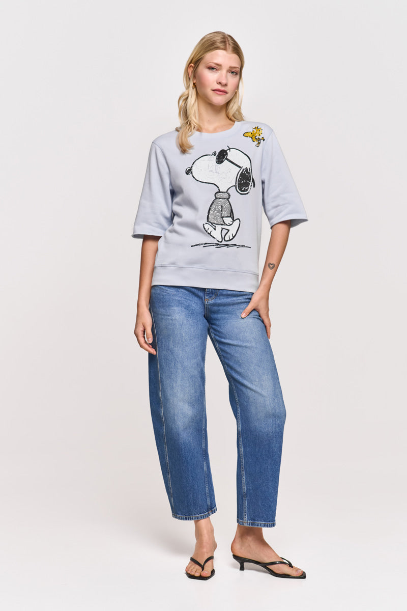 Eine Frau mit hellem Haar steht mit dem Gesicht nach vorne und trägt einen hellgrauen Baumwollmix-Sweatpullover mit Snoopy-Aufdruck, blaue Jeans und schwarze Flip-Flops. Ihr Haar ist offen, ihr Gesichtsausdruck neutral, und eine Hand steckt in ihrer Tasche.