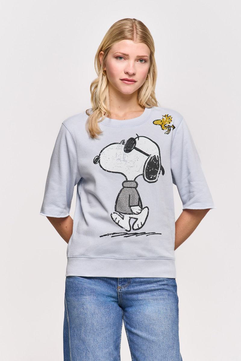 Eine junge blonde Frau steht mit dem Gesicht nach vorne, die Hände in den Taschen, und trägt eine blaue Jeans und einen hellblauen Princess Goes Hollywood-Pullover aus Baumwollmischgewebe mit einem großen Snoopy- und Woodstock-Aufdruck. Der Hintergrund ist einfarbig weiß.
