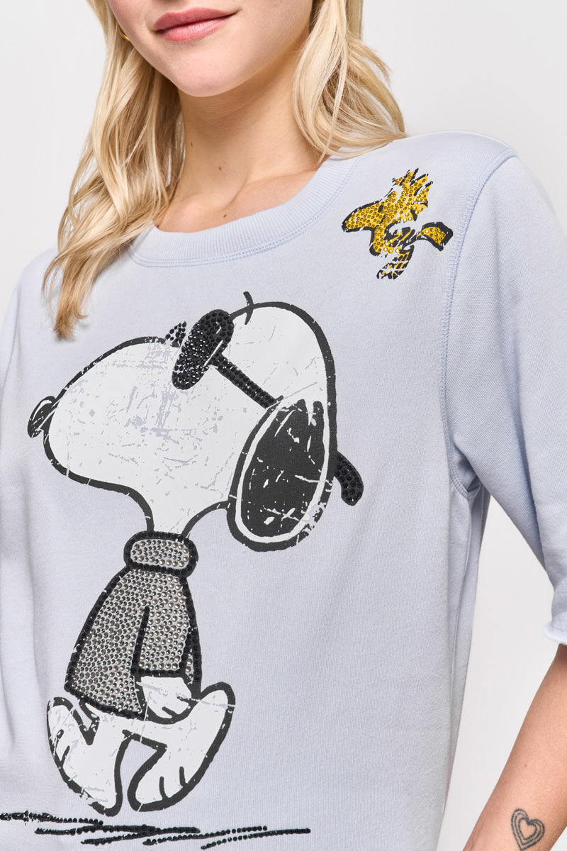 Eine Frau mit blonden Haaren trägt einen hellblauen Sweatpullover von Princess Goes Hollywood aus einem Baumwollgemisch mit einem Snoopy-Aufdruck mit Snoopy mit Sonnenbrille und Woodstock über ihm. Ihr lächelndes Gesicht und ein kleines Herz-Tattoo auf ihrem Unterarm sind zu sehen.