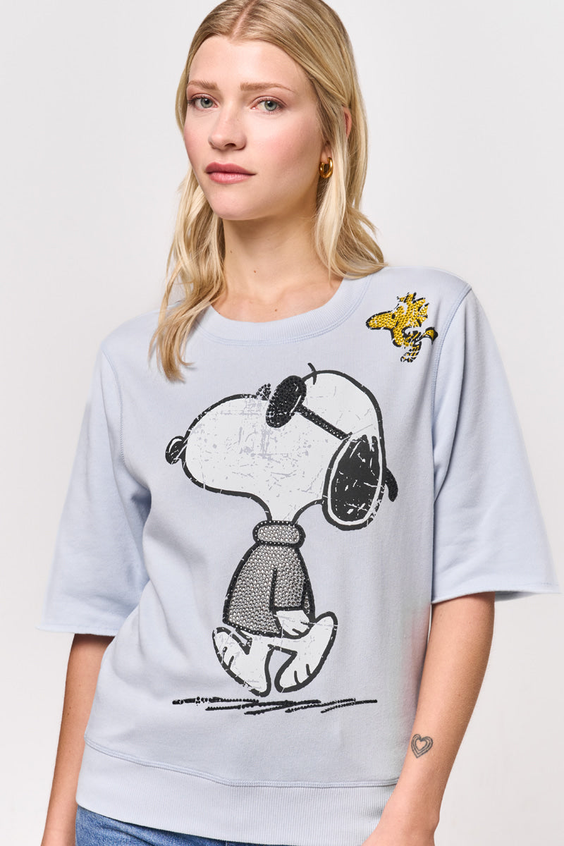 Eine Frau mit langen blonden Haaren trägt einen hellblauen Princess Goes Hollywood Baumwollmix-Sweatpullover mit großem Snoopy- und Woodstock-Aufdruck. Sie trägt goldene Ohrringe und ein Herz-Tattoo auf ihrem linken Unterarm und steht vor einem schlichten weißen Hintergrund.