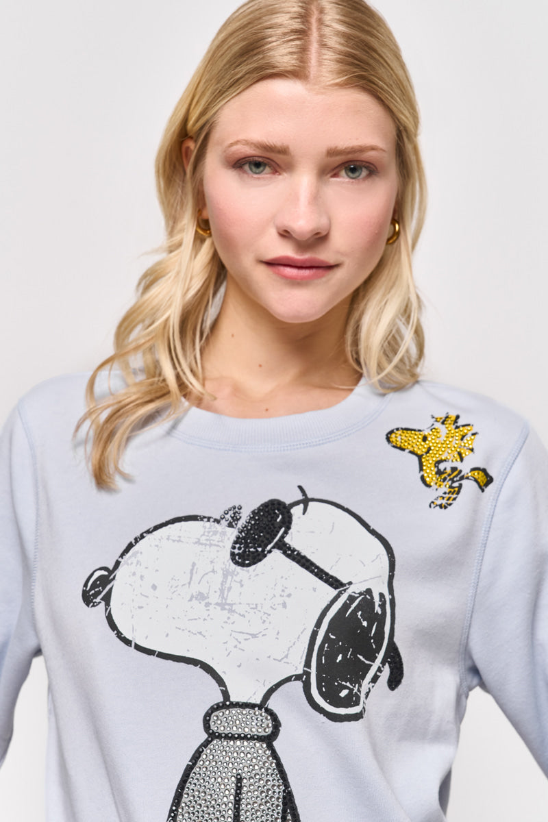 Eine blonde Frau trägt einen hellblauen Princess Goes Hollywood Sweatpullover aus Baumwollmix mit einem großen schwarz-weißen Snoopy-Aufdruck und einem kleinen gelben Woodstock in der Nähe ihrer linken Schulter. Sie steht mit neutralem Gesichtsausdruck vor einem schlichten Hintergrund.