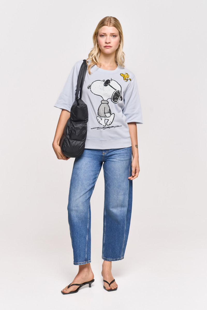 Eine Frau mit blonden Haaren trägt einen hellgrauen Princess Goes Hollywood Snoopy-Print Sweatpullover aus Baumwollmix, blaue Jeans und schwarze Flip-Flops. Sie trägt eine schwarze Umhängetasche und steht vor einem weißen Hintergrund und schaut neutral in die Kamera.