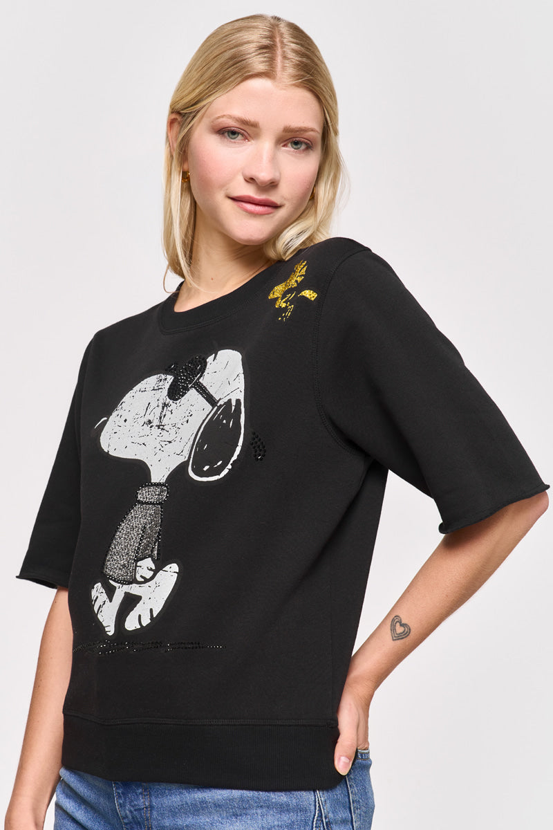 Eine blonde Frau mit glattem Haar trägt eine blaue Jeans und einen schwarzen Princess Goes Hollywood Baumwollmix-Sweatpullover mit einem Snoopy- und Woodstock-Aufdruck. Sie steht seitlich, lächelt sanft, legt die Hand auf die Hüfte und hat ein Herz-Tattoo auf ihrem linken Handgelenk.