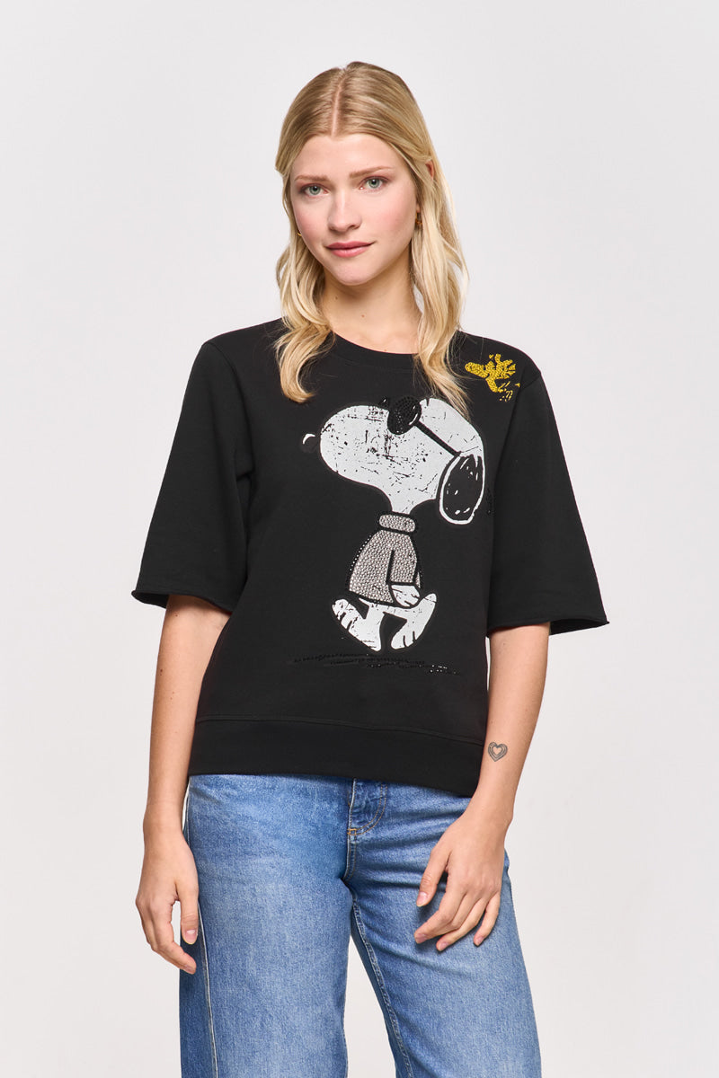 Eine blonde Frau trägt einen schwarzen Baumwollmix-Snoopy-Print-Sweatpullover von Princess Goes Hollywood mit einem gelben Woodstock auf der Schulter. Sie blickt in blauen Jeans mit neutralem Gesichtsausdruck nach vorne und hat ein Herz-Tattoo auf dem linken Unterarm.