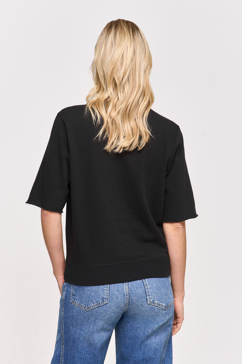 Eine Person mit gewelltem, blondem Haar trägt einen Princess Goes Hollywood Snoopy-Print Sweatpullover aus einer weichen Baumwollmischung und blaue Jeans und steht mit dem Rücken zur Kamera vor einem schlichten, hellen Hintergrund.