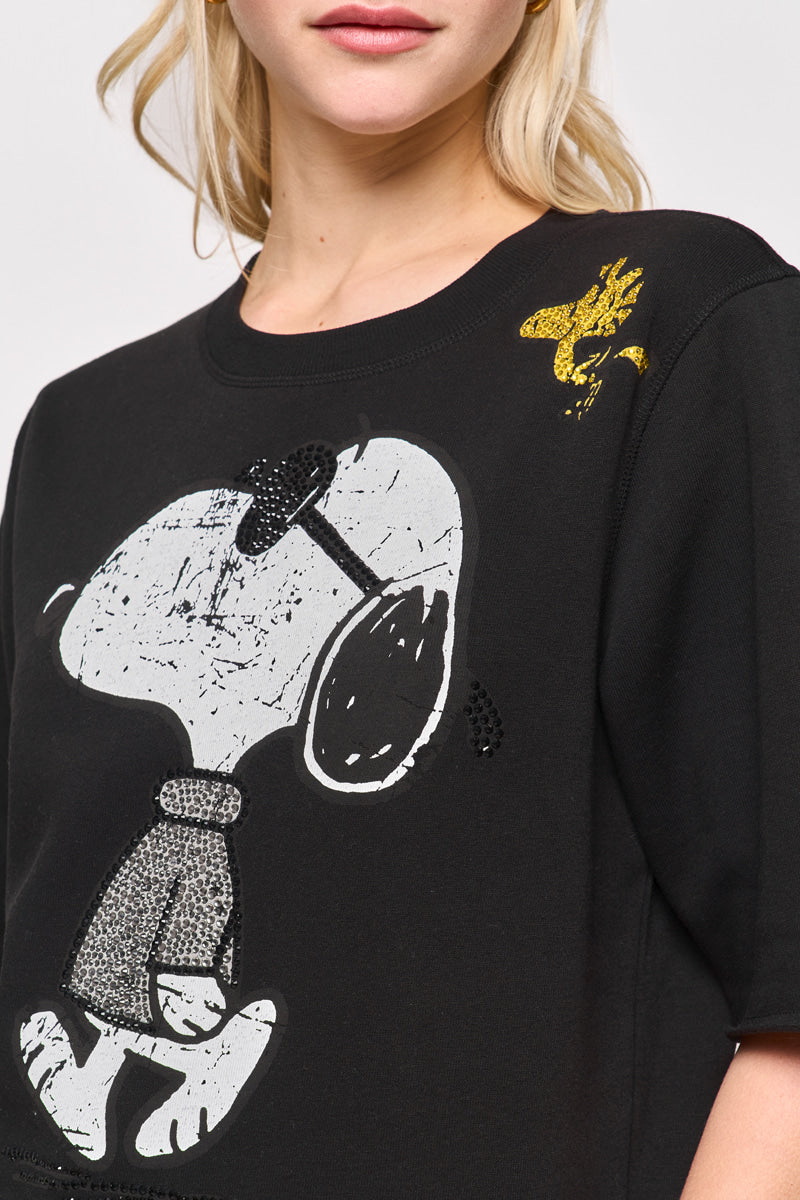Eine Frau trägt ein schwarzes Princess Goes Hollywood Baumwollmix-T-Shirt mit einem großen weißen Snoopy-Aufdruck in der Sonnenbrille. Eine kleine gelbe Woodstock-Grafik befindet sich auf der rechten Schulter. Nur ihr unteres Gesicht, ihr Hals und ihr oberer Oberkörper sind zu sehen.