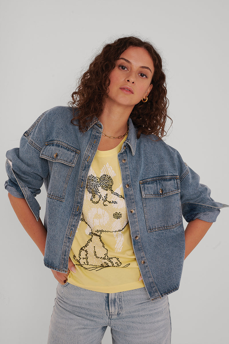 Eine Frau mit lockigen braunen Haaren trägt eine Princess Goes Hollywood Damen Jeansjacke über einem gelben, glitzernden Snoopy-T-Shirt, gepaart mit hellblauen Jeans. Sie steht selbstbewusst vor einem schlichten hellen Hintergrund.
