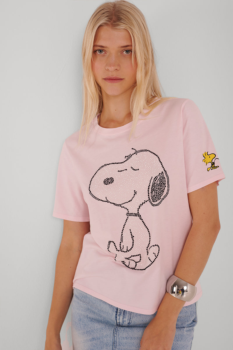 Eine Frau mit langen blonden Haaren trägt ein hellrosafarbenes Princess Goes Hollywood Snoopy T-Shirt aus Baumwolle mit einer großen umrandeten Snoopy-Grafik und einem kleinen Woodstock auf dem Ärmel, gepaart mit hellblauen Jeans und einem klobigen weißen Armband.