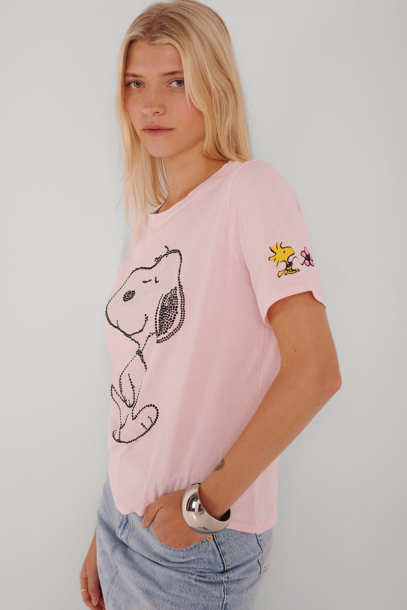 Eine junge Frau mit langen blonden Haaren trägt ein hellrosa Princess Goes Hollywood Snoopy T-Shirt aus Baumwolle mit einem großen gepunkteten Snoopy-Umriss auf der Vorderseite und einem kleinen Woodstock auf dem Ärmel, gepaart mit hellblauen Jeans-Shorts und einem silbernen Manschetten-Armband.