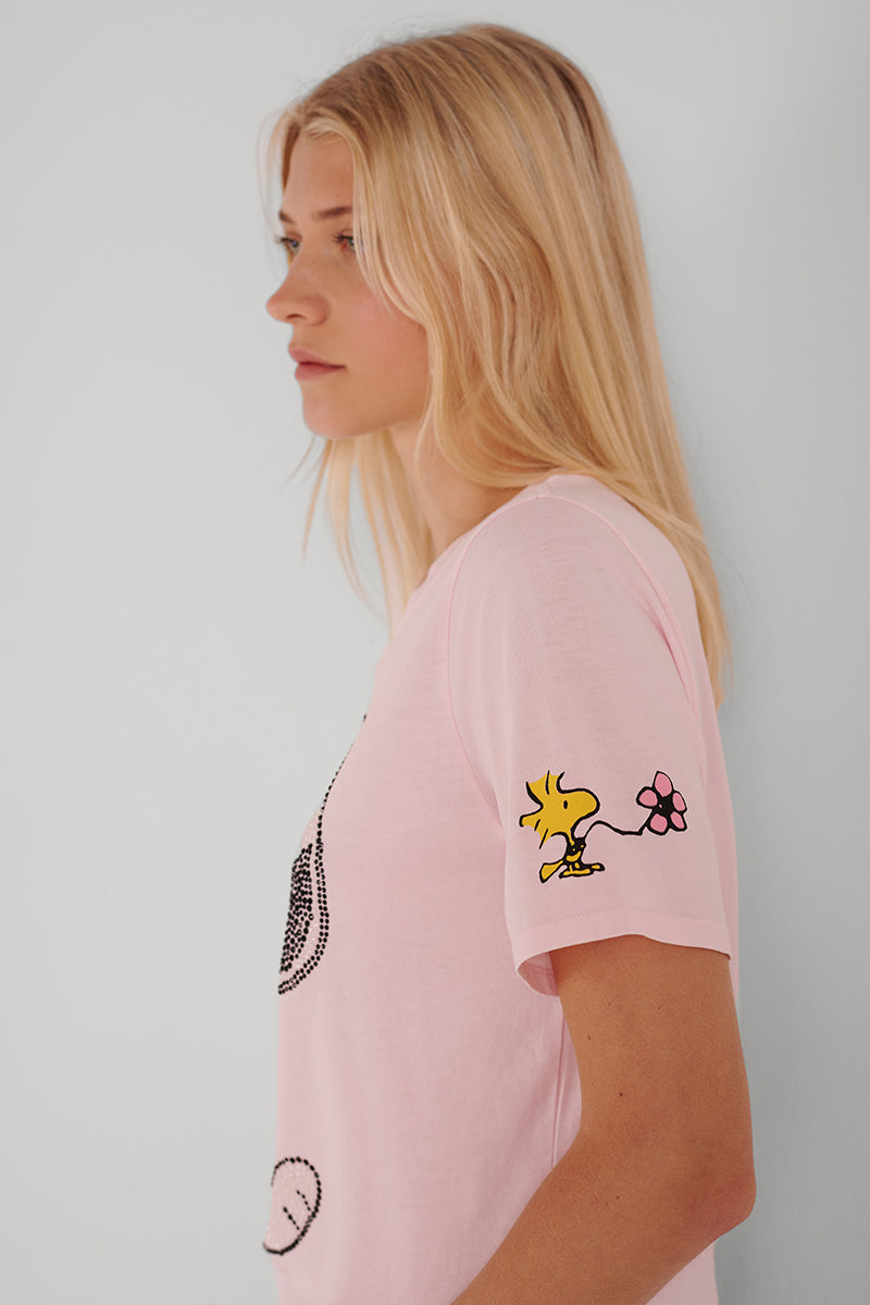 Eine Frau mit langen blonden Haaren trägt ein hellrosa Princess Goes Hollywood Baumwolle Snoopy T-Shirt, abgebildet im Profil vor einem weißen Hintergrund. Auf dem T-Shirt ist Woodstock abgebildet, der eine rosa Blume auf dem Ärmel hält. Ihr Gesichtsausdruck ist neutral.