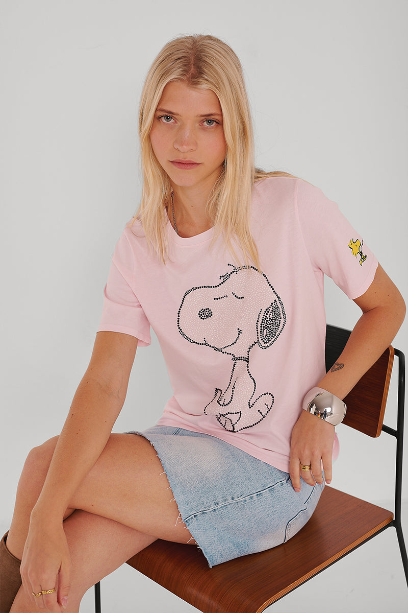 Eine Frau mit langen blonden Haaren sitzt auf einem Holzstuhl und trägt ein hellrosa Princess Goes Hollywood Snoopy-Baumwoll-T-Shirt mit einem großen schwarzen Snoopy-Umriss und einem kleinen Woodstock auf dem Ärmel, dazu einen hellblauen Jeansrock und ein klobiges Silberarmband.