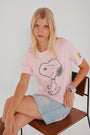 Eine Frau mit langen blonden Haaren sitzt auf einem Holzstuhl und trägt ein hellrosa Princess Goes Hollywood Snoopy-Baumwoll-T-Shirt mit einem großen schwarzen Snoopy-Umriss und einem kleinen Woodstock auf dem Ärmel, dazu einen hellblauen Jeansrock und ein klobiges Silberarmband.