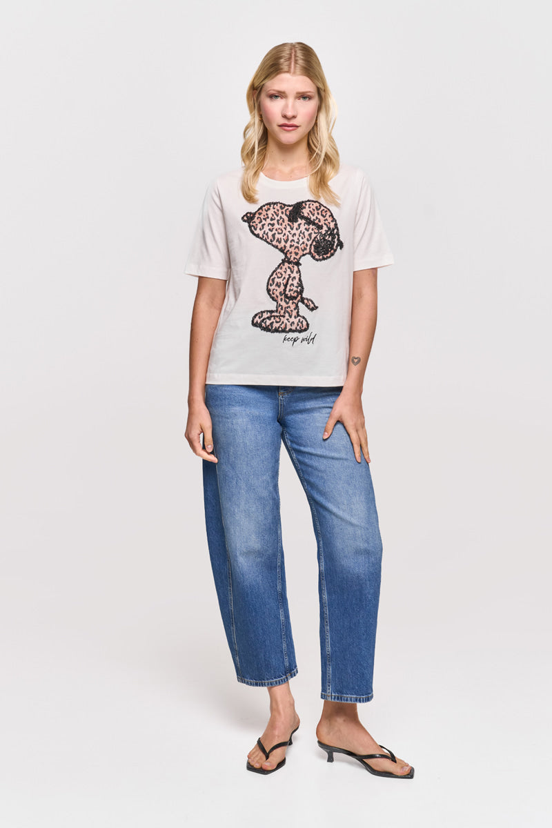 Eine blonde Frau steht vor einem schlichten weißen Hintergrund und trägt ein weißes Snoopy-T-Shirt von Princess Goes Hollywood mit einer großen Snoopy-Grafik mit Leopardenaufdruck und dem Text "Snoopy cool", weite Bluejeans und schwarze hochhackige Sandalen. Sie hat einen neutralen Gesichtsausdruck.