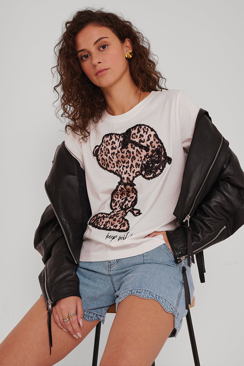 Eine Frau mit lockigen braunen Haaren sitzt auf einem Hocker und trägt ein weißes Princess Goes Hollywood-T-Shirt mit einer Snoopy-Grafik im Leopardenmuster, elastische Jeanshorts mit Bindebändern und eine schwarze Lederjacke über den Schultern. Sie blickt selbstbewusst in die Kamera.