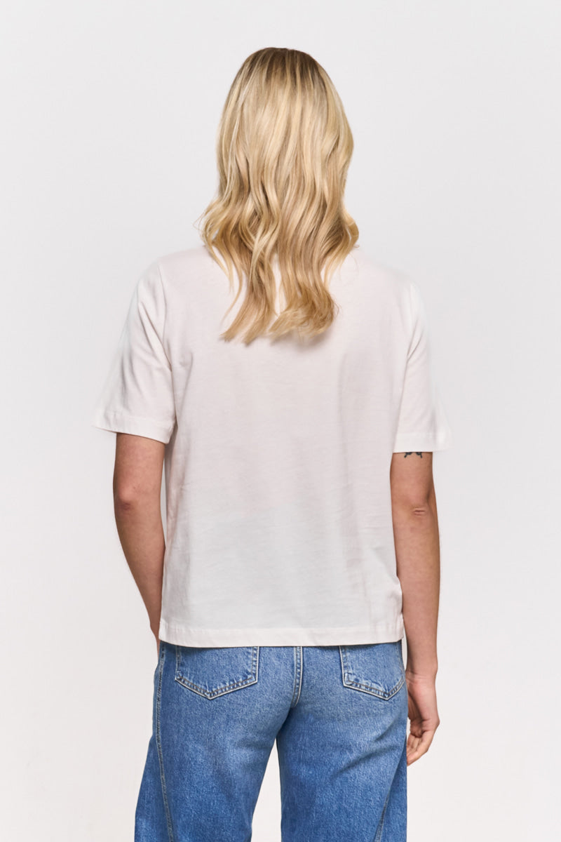 Eine Person mit schulterlangem blondem Haar blickt nach vorne und trägt ein schlichtes weißes Snoopy-T-Shirt von Princess Goes Hollywood und blaue Jeans mit sichtbaren Gesäßtaschen. Ihre entspannte Pose wird durch einen einfarbigen weißen Hintergrund hervorgehoben.