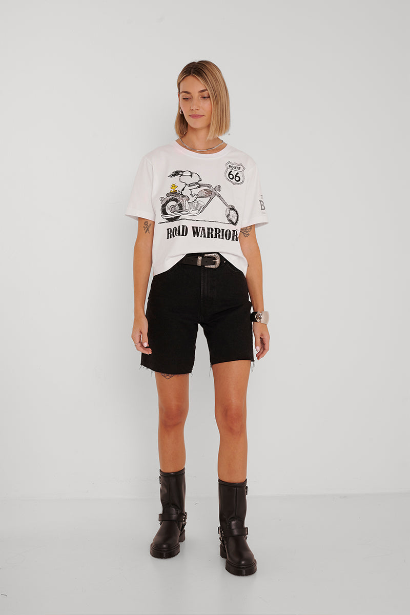 Eine Frau mit blondem Bubikopf steht vor einem weißen Hintergrund und trägt ein übergroßes T-Shirt mit dem Aufdruck Princess Goes Hollywood Snoopy, schwarze Shorts mit Gürtel und schwarze halbhohe Stiefel. Sie hat die Hände an den Seiten und einen neutralen Gesichtsausdruck.