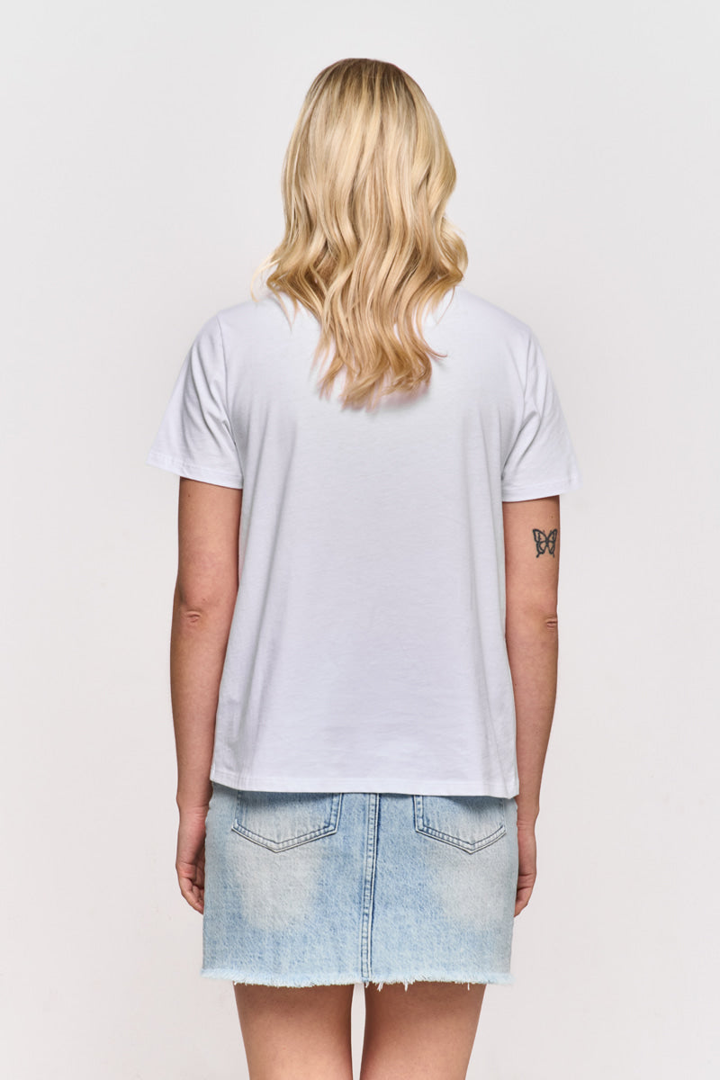 Eine Person mit langem, gewelltem blondem Haar steht mit dem Rücken zur Kamera, trägt ein weißes Princess Goes Hollywood Baumwolle T-Shirt und einen hellblauen Jeansrock. Auf ihrem linken Oberarm ist ein Schmetterlings-Tattoo vor einem schlichten weißen Hintergrund zu sehen.