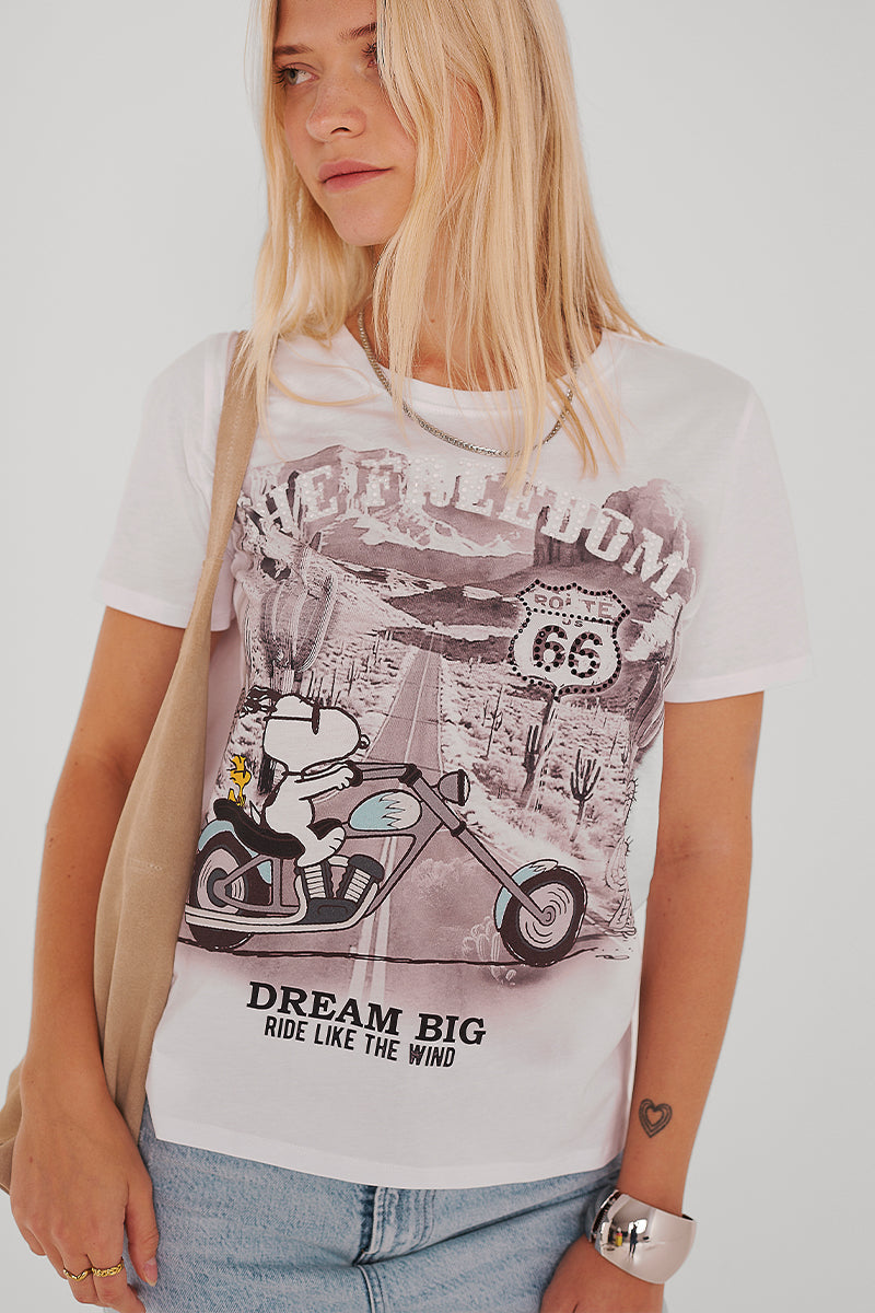 Eine Frau mit langen blonden Haaren trägt ein Princess Goes Hollywood T-Shirt mit Snoopy-Print aus Baumwolle, das Snoopy auf einem Motorrad und den Slogan "DREAM BIG, RIDE LIKE THE WIND" zeigt. Sie hat außerdem ein Herz-Tattoo am Handgelenk und trägt eine beigefarbene Tasche.