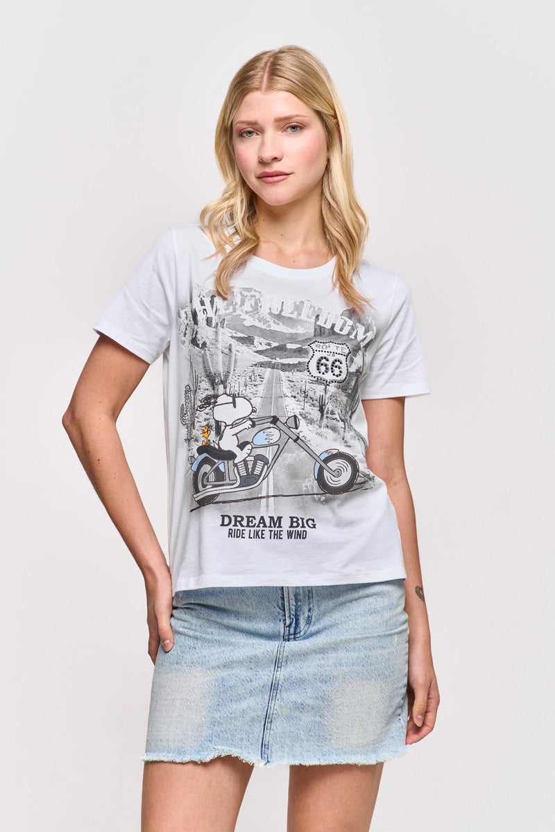 Eine junge Frau mit langen blonden Haaren trägt ein weißes Baumwoll-T-Shirt von Princess Goes Hollywood mit einem Cartoon-Hund auf einem Motorrad und dem Text "Dream Big, Ride Like The Wind", gepaart mit einem hellblauen Jeansminirock vor einem schlichten weißen Hintergrund.