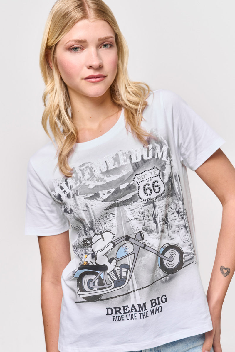 Eine junge Frau mit langen blonden Haaren trägt ein weißes Baumwoll-T-Shirt von Princess Goes Hollywood, auf dem ein Snoopy auf einem Motorrad, Route 66-Zeichen und der Slogan "DREAM BIG RIDE LIKE THE WIND" abgebildet sind. Sie lächelt sanft vor einem schlichten hellen Hintergrund.
