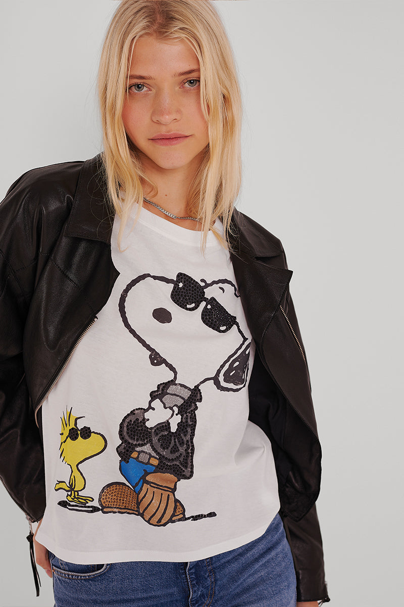 Eine blonde Frau trägt eine schwarze Lederjacke über einem Princess Goes Hollywood Snoopy T-Shirt aus Baumwolle, mit Snoopy mit Sonnenbrille und Woodstock, gepaart mit blauen Jeans. Sie steht mit dem Gesicht nach vorne vor einem schlichten hellen Hintergrund.