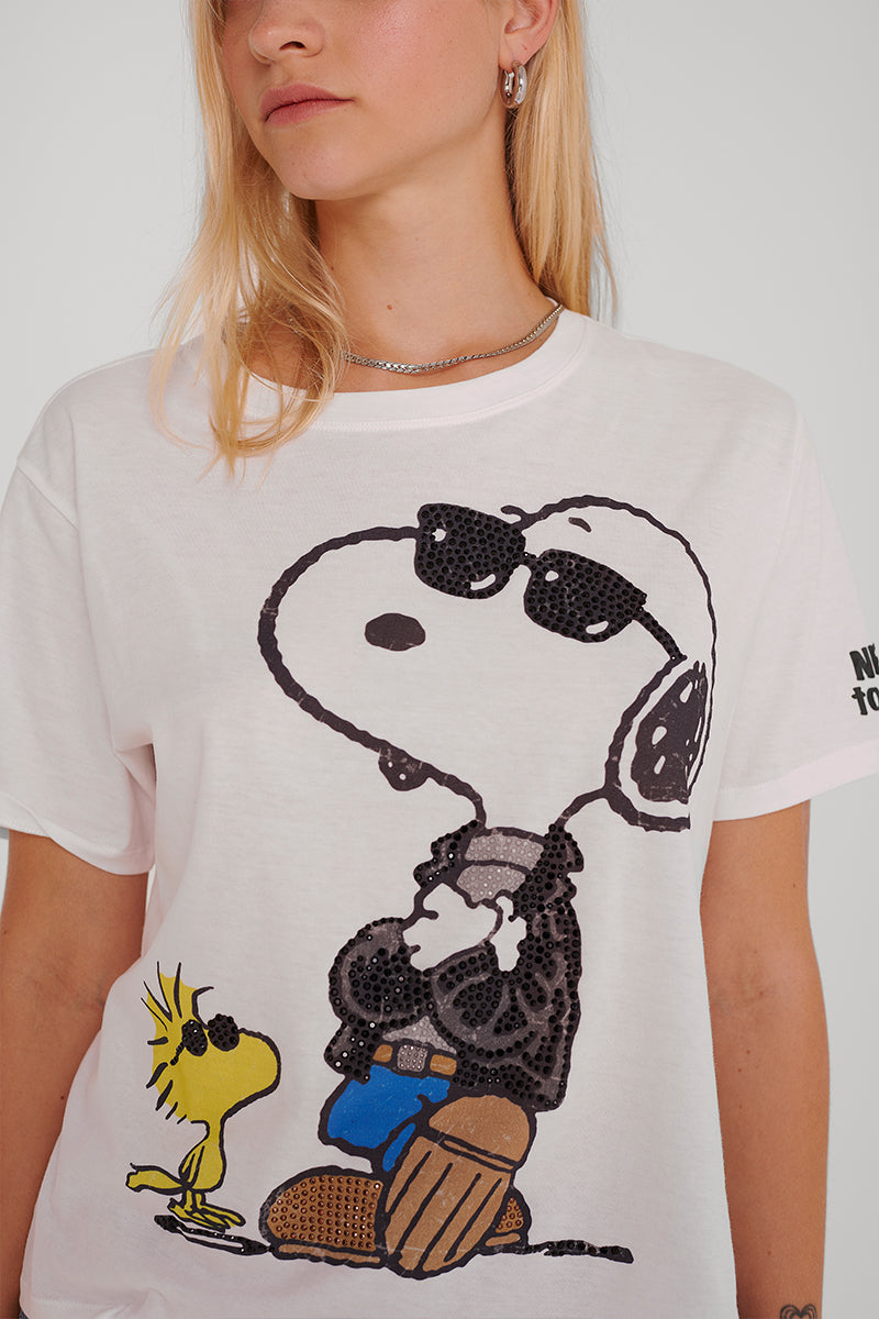 Eine Person mit langen blonden Haaren trägt ein Princess Goes Hollywood Snoopy-T-Shirt, das Snoopy mit Sonnenbrille, Lederjacke, Handschuhen und Blue Jeans mit verschränkten Armen zeigt. Woodstock steht neben ihm auf einem einfarbigen Hintergrund.