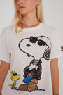 Eine Person mit langen blonden Haaren trägt ein Princess Goes Hollywood Snoopy-T-Shirt, das Snoopy mit Sonnenbrille, Lederjacke, Handschuhen und Blue Jeans mit verschränkten Armen zeigt. Woodstock steht neben ihm auf einem einfarbigen Hintergrund.