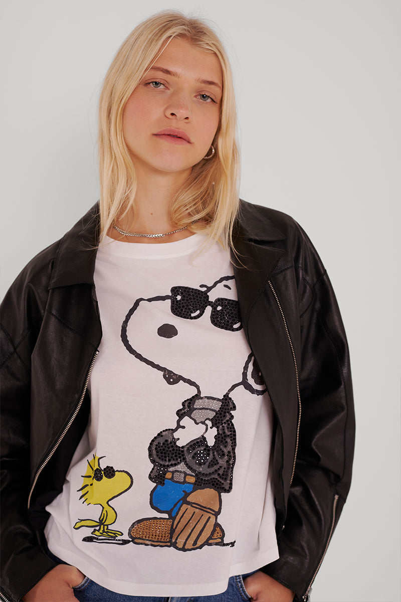Eine blonde Frau trägt eine schwarze Lederjacke über einem Princess Goes Hollywood Snoopy Baumwoll-T-Shirt, auf dem Woodstock mit Sonnenbrille abgebildet ist. Sie steht entspannt vor einem weißen Hintergrund, ihr Haar ist offen und ihre Hände stecken in den Taschen ihrer Jeans.