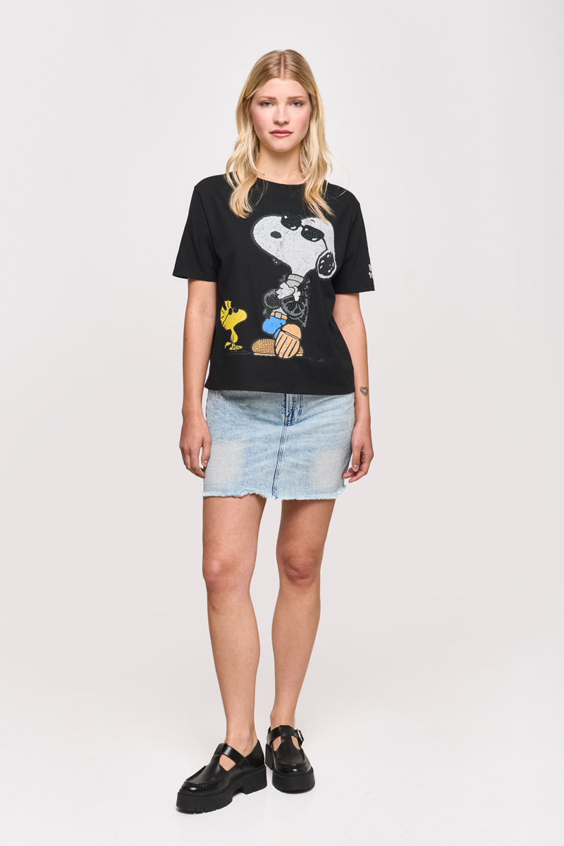 Eine Frau mit blondem Haar trägt ein schwarzes Princess Goes Hollywood Snoopy-T-Shirt mit Snoopy und Woodstock, einen hellblauen Jeansminirock und schwarze, klobige Schuhe. Sie steht mit dem Gesicht nach vorne vor einem schlichten weißen Hintergrund, die Arme locker an den Seiten.