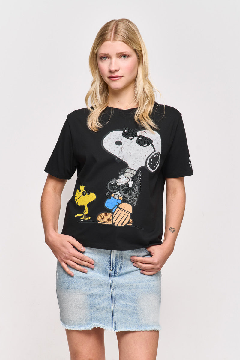 Eine junge Frau mit glattem blondem Haar trägt ein schwarzes Snoopy-T-Shirt aus Baumwolle von Princess Goes Hollywood mit Woodstock und einen hellblauen Denim-Rock. Sie steht vor einem weißen Hintergrund, die Arme locker in die Hüften gestemmt, mit einem neutralen Gesichtsausdruck.