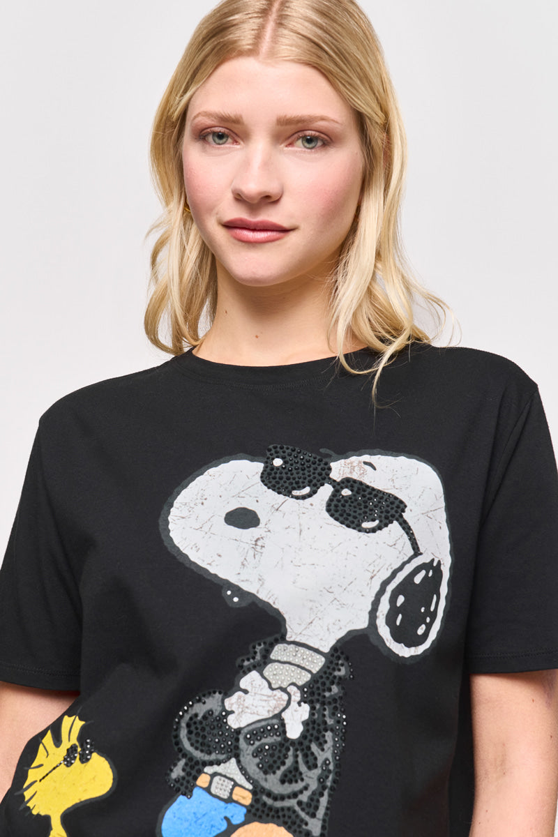 Eine junge blonde Frau trägt ein Princess Goes Hollywood Snoopy T-Shirt aus Baumwolle, das Snoopy mit Sonnenbrille und schwarzem Outfit mit Woodstock auf der linken Seite zeigt. Der kräftige Aufdruck hebt sich deutlich von dem schlichten hellen Hintergrund ab.