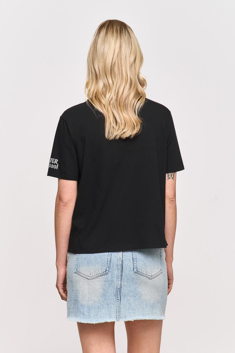 Eine Frau mit gewelltem blondem Haar blickt von sich weg und trägt ein schwarzes Princess Goes Hollywood Snoopy-T-Shirt und einen hellblauen Jeansrock mit sichtbaren Gesäßtaschen. Ihre Arme ruhen an ihren Seiten vor einem schlichten, hellen Hintergrund.