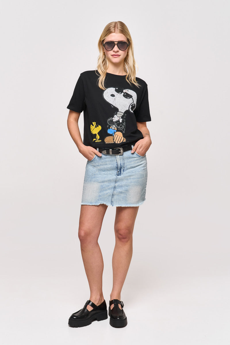 Eine blonde Frau trägt eine Sonnenbrille, ein schwarzes Princess Goes Hollywood Snoopy-T-Shirt mit Woodstock-Aufdruck, einen hellblauen Jeansminirock mit Strasssteinen und schwarze Plateauschuhe. Sie steht mit dem Gesicht nach vorne, eine Hand in der Rocktasche, vor einem schlichten Hintergrund.