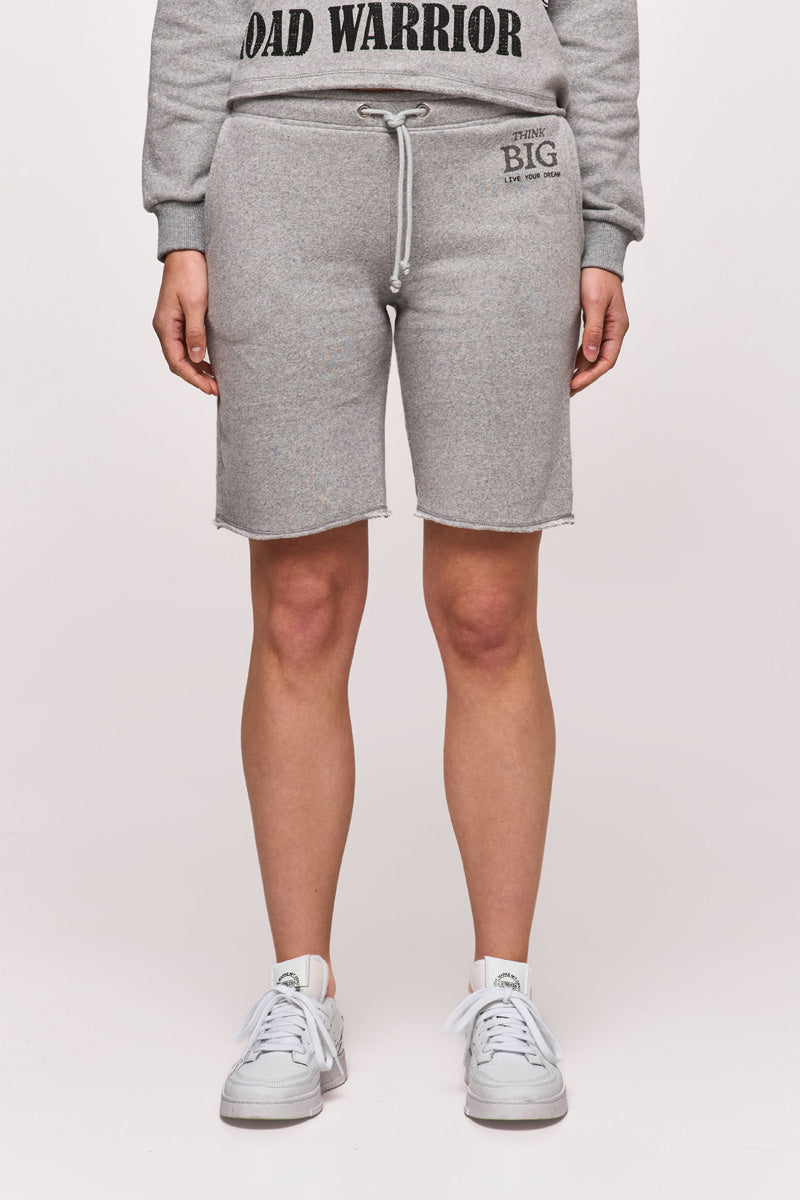 Eine nach vorne blickende Person in hellgrauen Princess Goes Hollywood Sweat-Bermudashorts mit elastischem Bund und THINK BIG Text auf dem linken Oberschenkel, einem passenden Sweatshirt mit teilweise sichtbarem ROAD WARRIOR und weißen Turnschuhen vor einem schlichten hellen Hintergrund.