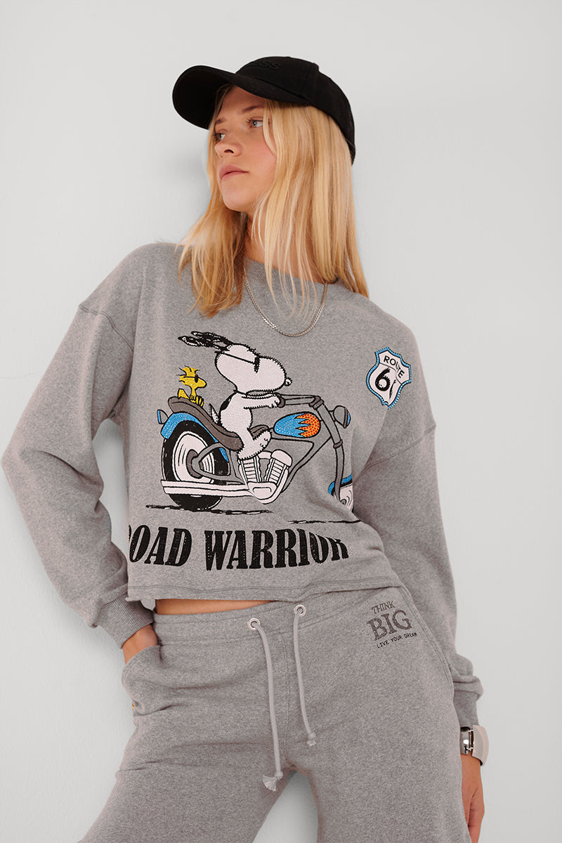 Eine junge Frau mit langen blonden Haaren trägt eine schwarze Baseballkappe, eine graue Jogginghose mit Kordelzug und ein graues Sweatshirt in Übergröße mit dem Aufdruck eines Snoopy-Motorrads ROAD WARRIOR und steht selbstbewusst vor einer schlichten Wand.