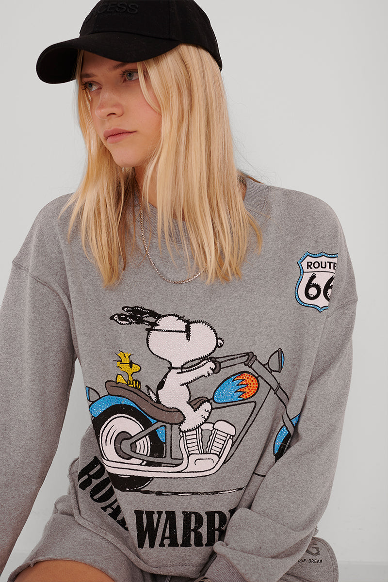 Eine Frau mit langen blonden Haaren trägt eine schwarze Mütze und einen übergroßen grauen Princess Goes Hollywood-Sweatpullover mit Snoopy und Woodstock auf einem Motorrad, einem Route 66-Aufnäher und "Road Warrior". Sie blickt leicht zur Seite, ihr Blick ist neutral.
