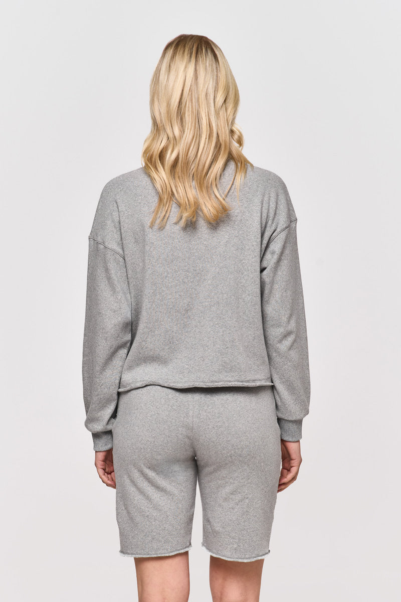 Eine Person mit langem, gewelltem blondem Haar steht mit dem Gesicht zur Wand und trägt Princess Goes Hollywood hellgraue Sweat-Bermudashorts mit elastischem Bund und passendem Sweatshirt. Der schlichte weiße Hintergrund hebt das lässige Baumwoll-Outfit hervor.