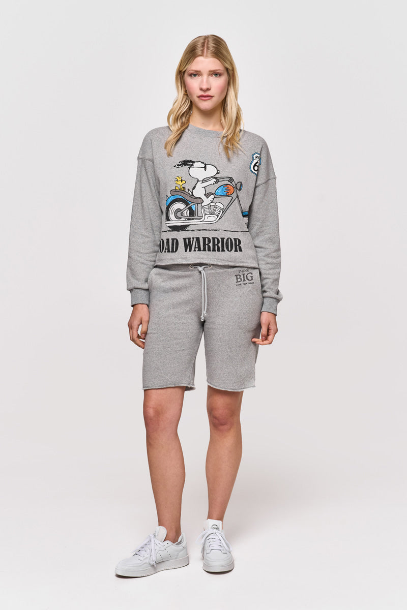 Eine blonde Frau blickt nach vorne in einem Princess Goes Hollywood Sweatpullover mit Snoopy-Print und der Aufschrift "ROAD WARRIOR", gepaart mit passenden grauen Shorts und weißen Turnschuhen, vor einem schlichten weißen Hintergrund.