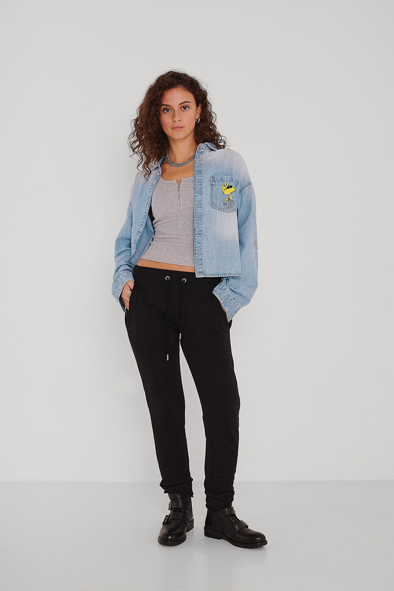 Eine Person mit lockigem Haar steht vor einer weißen Wand. Sie trägt ein hellblaues Princess Goes Hollywood-Jeanshemd mit einem gelben Woodstock-Aufnäher, ein graues Crop-Top, eine schwarze Jogginghose und schwarze Ankle Boots. Die Hände sind in den Taschen; der Gesichtsausdruck ist neutral.