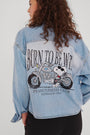 Eine Person mit lockigem braunem Haar, von hinten gesehen, trägt ein hellblaues Princess Goes Hollywood Jeanshemd mit einem Peanuts-Snoopy-Motorradaufdruck. "BORN TO BE WILD" steht über "PEANUTS BIKERS CLUB FOR BEAGLES ONLY".