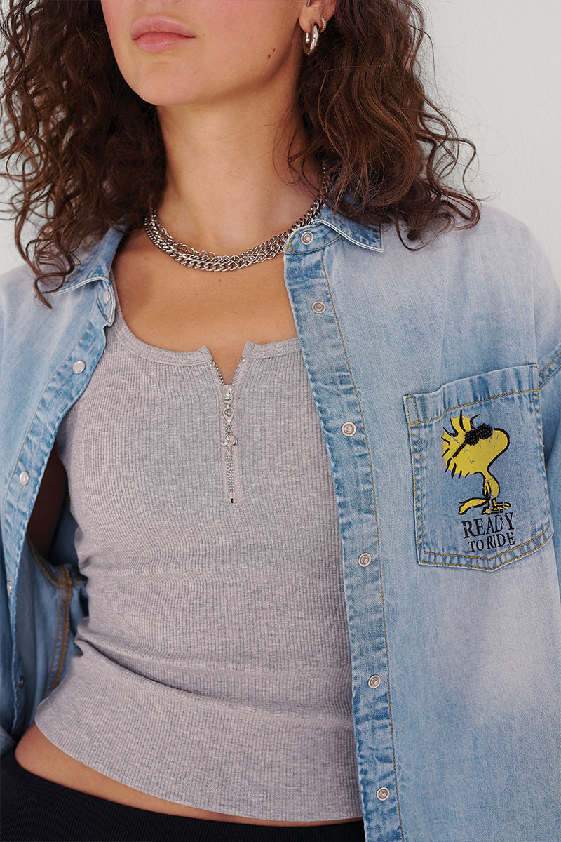 Eine Frau mit lockigem braunem Haar trägt mehrlagige Ketten, ein hellgraues geripptes Tank-Top mit Reißverschluss und ein offenes Princess Goes Hollywood Jeanshemd mit einem Snoopy "Ready to Ride!"-Aufnäher auf der Tasche. Nur ihr Oberkörper und ihr unteres Gesicht sind zu sehen.
