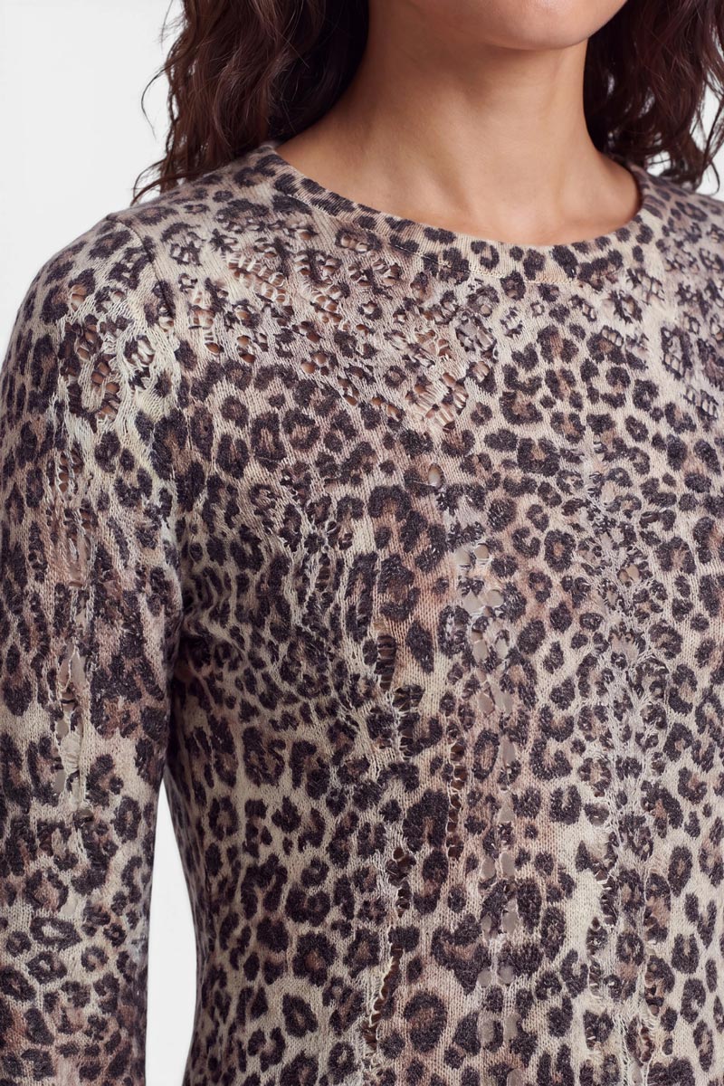 Eine Person mit welligem braunem Haar trägt einen eng anliegenden, langärmeligen Strickpullover mit auffälligem Leopardenmuster. Das Oberteil weist sichtbare Used-Effekte und Löcher auf, die auf eine einzigartige Textur aus einer Woll-Alpaka-Mischung hindeuten. Das Bild ist ab der Mitte der Brust nach oben abgeschnitten; der Hintergrund ist einfarbig weiß.
